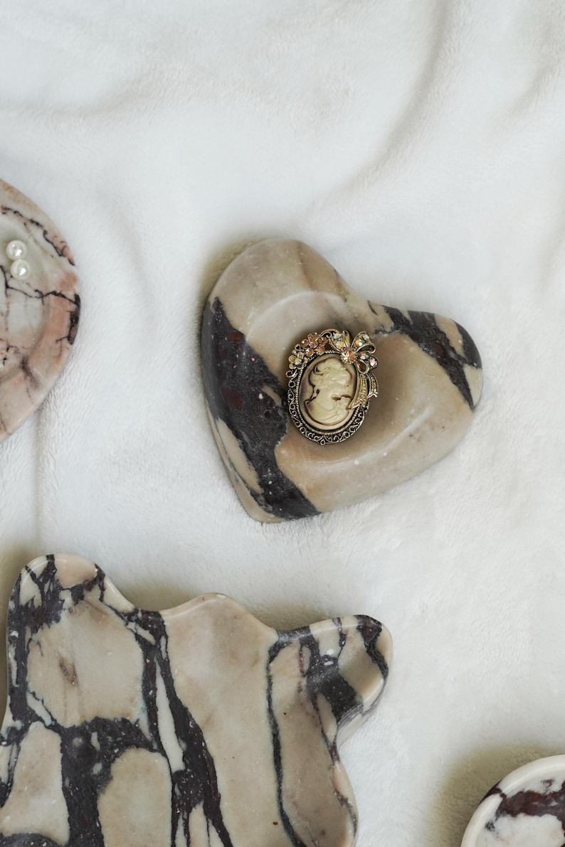 May include: An oval-shaped cameo brooch featuring a profile portrait in cream and brown tones, set within an ornate, antique-gold colored metal frame decorated with small floral accents and a bow detail at the top, resting on a heart-shaped stone.