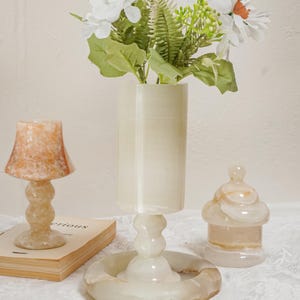 Puede incluir: Un conjunto de objetos decorativos de piedra transl&uacute;cida crema, que incluye una peque&ntilde;a l&aacute;mpara, un jarr&oacute;n con flores y un tarro con tapa. La l&aacute;mpara tiene una pantalla acampanada, y el jarr&oacute;n contiene flores blancas y vegetaci&oacute;n.