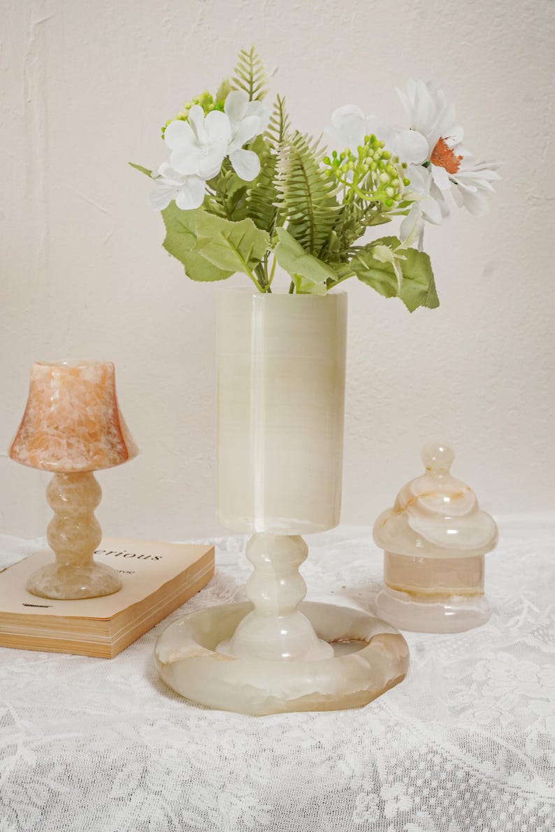 Puede incluir: Un conjunto de objetos decorativos de piedra transl&uacute;cida crema, que incluye una peque&ntilde;a l&aacute;mpara, un jarr&oacute;n con flores y un tarro con tapa. La l&aacute;mpara tiene una pantalla acampanada, y el jarr&oacute;n contiene flores blancas y vegetaci&oacute;n.