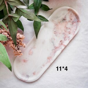 May include: A white and pink marble tray with rounded edges, measuring 11 x 4 inches. The tray has a marbled pattern with pink and red veining. The tray is displayed on a white surface, with greenery and pink flowers in the background.