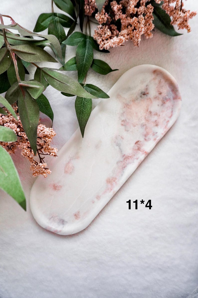May include: A white and pink marble tray with rounded edges, measuring 11 x 4 inches. The tray has a marbled pattern with pink and red veining. The tray is displayed on a white surface, with greenery and pink flowers in the background.