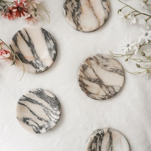 May include: A set of four circular coasters made of polished stone, featuring a cream-colored base with prominent dark grey and black veining patterns throughout each piece, arranged in a scattered formation alongside small white and pink flowers.