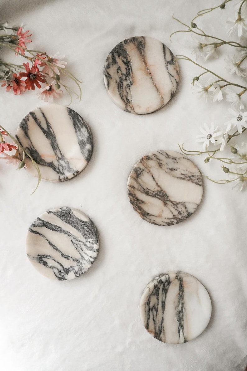 May include: A set of four circular coasters made of polished stone, featuring a cream-colored base with prominent dark grey and black veining patterns throughout each piece, arranged in a scattered formation alongside small white and pink flowers.