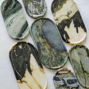 May include: A collection of seven oval-shaped stone trays featuring various natural patterns in shades of forest green, cream, black, and grey. Each tray has a smooth, polished surface with distinct veining and unique mineral color variations.