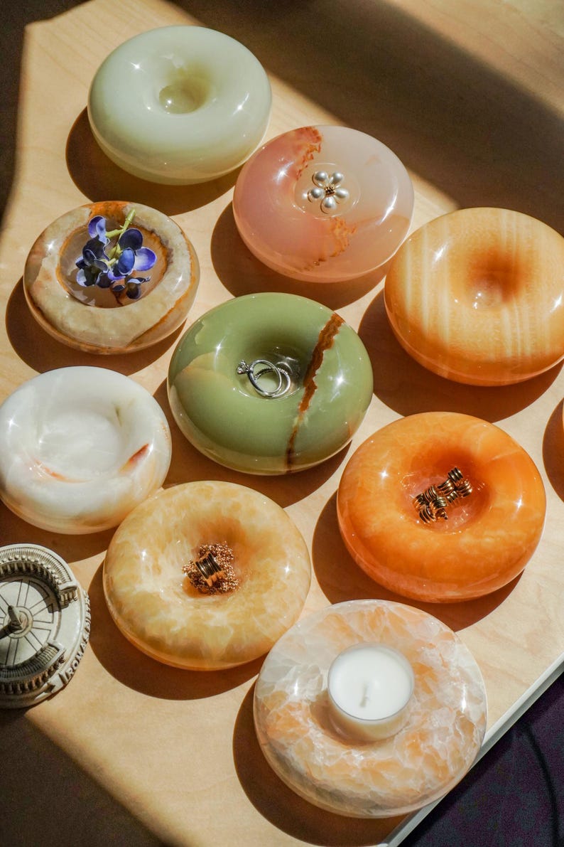 May include: A collection of smooth, donut-shaped stone trinket dishes in various colors including pale green, orange, and white. Some dishes hold small items like rings, a tea light candle, or a small blue flower, while others remain empty.