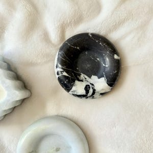 May include: A circular black marble dish with prominent white veining patterns throughout the stone surface. The dish features a smooth, polished finish and a concave center, presented alongside a smaller, solid light gray stone dish.