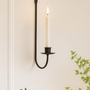 May include: A black metal wall sconce with a lit white candle. The sconce has a long, slender arm and a circular base to hold the candle. The candle is tall and cylindrical, with a visible flame. Decorative home decor.