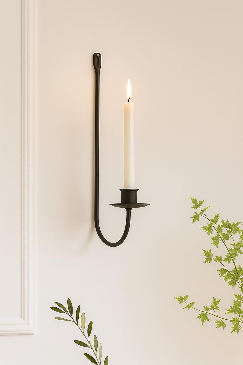 May include: A black metal wall sconce with a lit white candle. The sconce has a long, slender arm and a circular base to hold the candle. The candle is tall and cylindrical, with a visible flame. Decorative home decor.