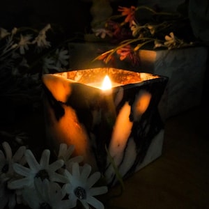 May include: A lit candle in a square marble container with black and white veining. The candle's flame glows brightly, illuminating the interior. White flowers and other decorative elements are in the background, creating a warm, inviting ambiance.