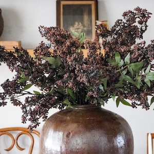 May include: A large, round, dark brown glazed ceramic vase with a wide body and a narrow neck. The vase is filled with a dense arrangement of dried, dark reddish-brown flowering branches interspersed with small, pointed green leaves.