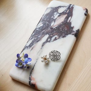 May include: A rectangular white marble tray with prominent dark purple and black veining. Resting on the surface are a small cluster of blue artificial flowers, a white faux pearl flower brooch, and a circular silver-toned filigree brooch.
