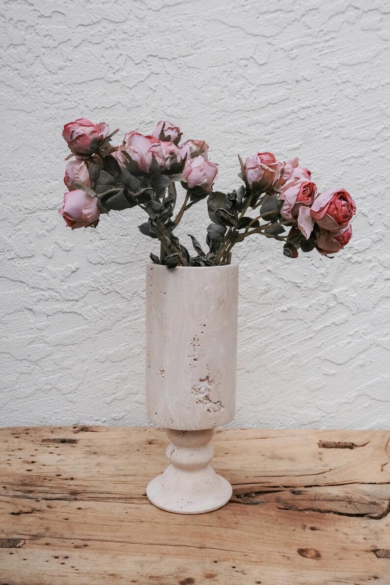 Puede incluir: Un jarr&oacute;n cil&iacute;ndrico rosa claro con base de pedestal contiene un ramo de rosas rosas secas. El jarr&oacute;n tiene un aspecto texturizado y desgastado. El arreglo se encuentra sobre una superficie de madera r&uacute;stica contra una pared blanca texturizada.