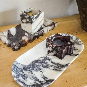 May include: Two marble candle holders and trays. One tray is oval, the other is rectangular with a scalloped edge. The candle holders are cube-shaped and star-shaped. The marble has a white base with dark gray and burgundy veining.