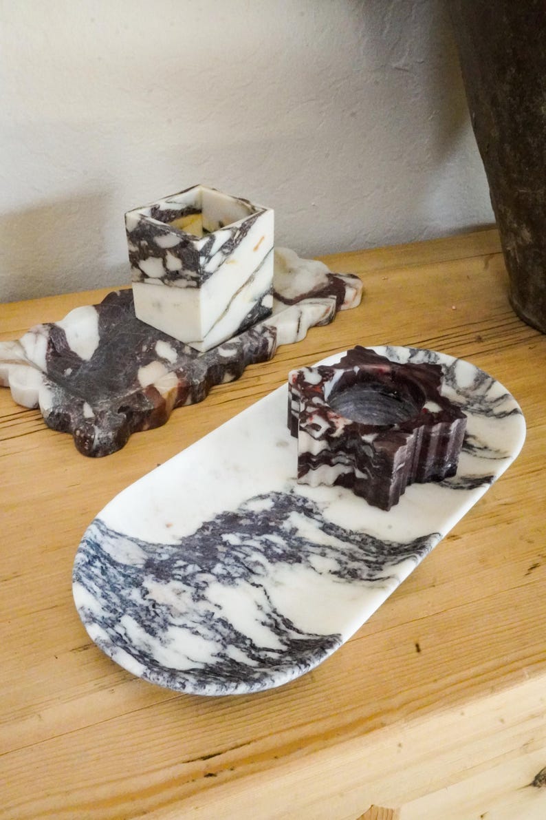 May include: Two marble candle holders and trays. One tray is oval, the other is rectangular with a scalloped edge. The candle holders are cube-shaped and star-shaped. The marble has a white base with dark gray and burgundy veining.