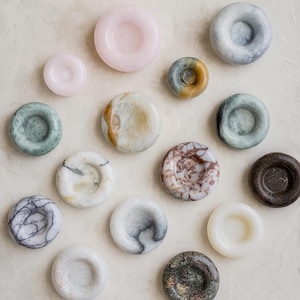 May include: A collection of various stone coasters in shades of pink, green, brown, and white. The coasters have a circular shape with a recessed center. The arrangement is on a neutral, textured surface.