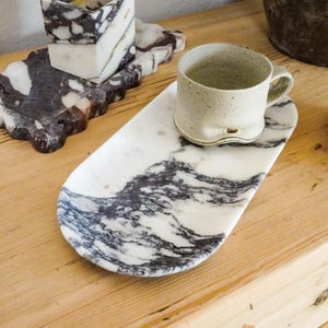 May include: A marble tray with a white and gray swirling pattern holds a beige ceramic mug. A square marble candle holder and a smaller marble tray with a similar pattern are also visible. The items are on a wooden surface.