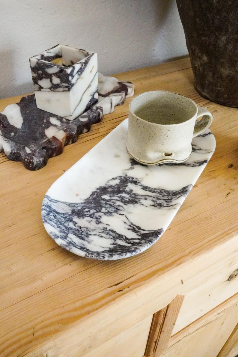 May include: A marble tray with a white and gray swirling pattern holds a beige ceramic mug. A square marble candle holder and a smaller marble tray with a similar pattern are also visible. The items are on a wooden surface.