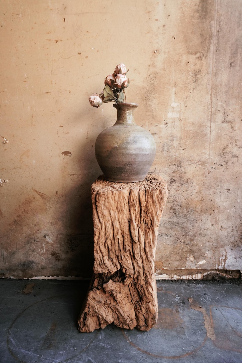 K&ouml;nnte beinhalten: Eine runde, erdfarbene Keramikvase mit engem Hals, in der sich ein kleines B&uuml;ndel getrockneter, blassrosa Blumen befindet. Die Vase steht auf einem hohen, verwitterten, vertikalen St&uuml;ck Treibholz mit einer rauen, strukturierten Oberfl&auml;che.