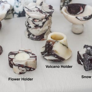 Puede incluir: Una colecci&oacute;n de portavelas y recipientes de m&aacute;rmol con patrones veteados en blanco y morado oscuro. El conjunto incluye un portavelas de columna, un tarro con tapa, un cuenco, un soporte con forma de flor, uno de volc&aacute;n y uno de estrella.