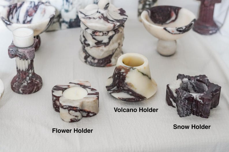 Puede incluir: Una colecci&oacute;n de portavelas y recipientes de m&aacute;rmol con patrones veteados en blanco y morado oscuro. El conjunto incluye un portavelas de columna, un tarro con tapa, un cuenco, un soporte con forma de flor, uno de volc&aacute;n y uno de estrella.