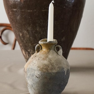 Puede incluir: Un pequeño jarrón de cerámica desgastada con una vela blanca. El jarrón tiene una combinación de colores de dos tonos, con una parte superior marrón claro y una parte inferior gris azulado. Dos asas pequeñas están a cada lado del jarrón. Un jarrón grande de cerámica marrón oscuro en el fondo.