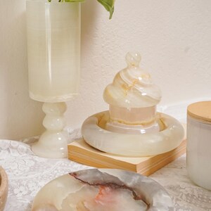 Puede incluir: Colecci&oacute;n de objetos decorativos de &oacute;nix, que incluye un jarr&oacute;n alto, un recipiente con tapa y un cenicero. Las piezas son en tonos crema, beige y marr&oacute;n, con una calidad transl&uacute;cida. Los objetos se exhiben sobre un mantel de encaje blanco.