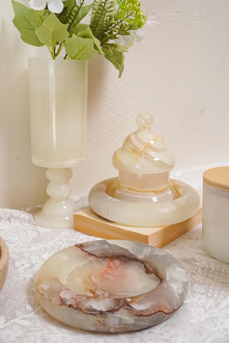 Puede incluir: Colecci&oacute;n de objetos decorativos de &oacute;nix, que incluye un jarr&oacute;n alto, un recipiente con tapa y un cenicero. Las piezas son en tonos crema, beige y marr&oacute;n, con una calidad transl&uacute;cida. Los objetos se exhiben sobre un mantel de encaje blanco.