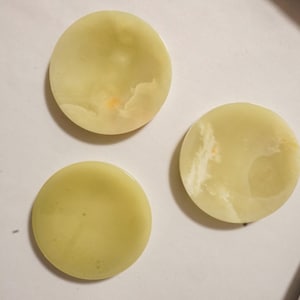 May include: Three circular, pale green stone coasters with smooth, polished surfaces. Each piece displays natural, subtle variations in color and cloudy, translucent patterns throughout the stone, creating a unique look for each individual item.