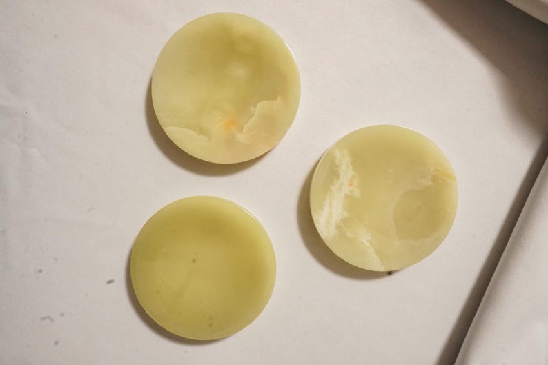 May include: Three circular, pale green stone coasters with smooth, polished surfaces. Each piece displays natural, subtle variations in color and cloudy, translucent patterns throughout the stone, creating a unique look for each individual item.