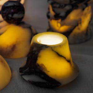 Puede incluir: Un portavelas en forma de campana hecho de piedra transl&uacute;cida con un patr&oacute;n veteado en tonos crema, amarillo y marr&oacute;n oscuro. La piedra est&aacute; iluminada desde el interior, emitiendo un brillo c&aacute;lido, con una abertura circular en la parte superior.
