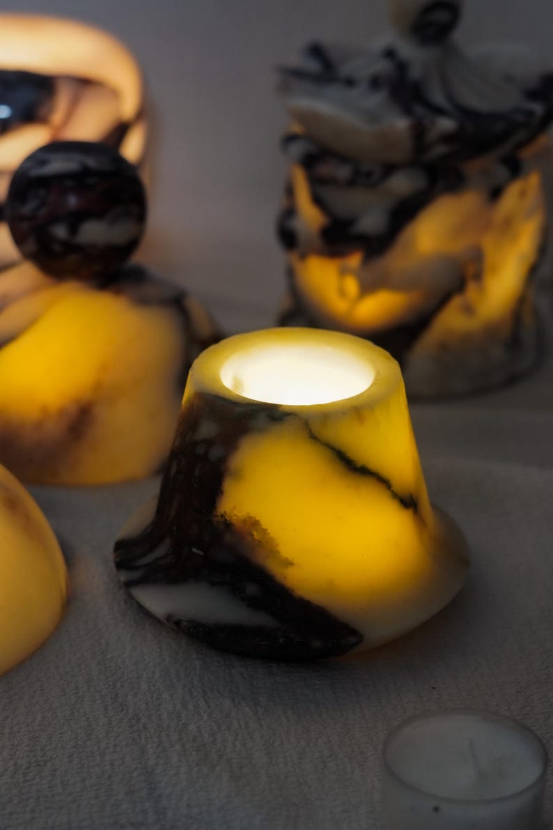 Puede incluir: Un portavelas en forma de campana hecho de piedra transl&uacute;cida con un patr&oacute;n veteado en tonos crema, amarillo y marr&oacute;n oscuro. La piedra est&aacute; iluminada desde el interior, emitiendo un brillo c&aacute;lido, con una abertura circular en la parte superior.
