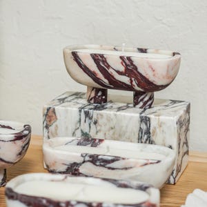 Puede incluir: Tres recipientes para velas de m&aacute;rmol de forma ovalada con cuatro patas cortas cada uno, con una base blanca con vetas de color rojo oscuro y negro. Cada recipiente contiene una vela de cera blanca con una mecha centrada. Un bloque rectangular est&aacute; detr&aacute;s.