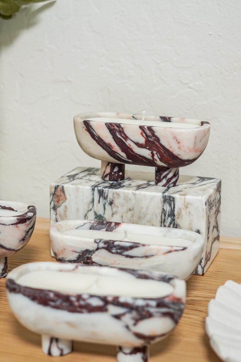 Puede incluir: Tres recipientes para velas de m&aacute;rmol de forma ovalada con cuatro patas cortas cada uno, con una base blanca con vetas de color rojo oscuro y negro. Cada recipiente contiene una vela de cera blanca con una mecha centrada. Un bloque rectangular est&aacute; detr&aacute;s.