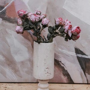 Puede incluir: Un jarr&oacute;n cil&iacute;ndrico blanco con base de pedestal, lleno de rosas rosas. El jarr&oacute;n tiene una superficie texturizada y est&aacute; sobre una superficie de madera. El fondo presenta una pintura abstracta con tonos neutros.