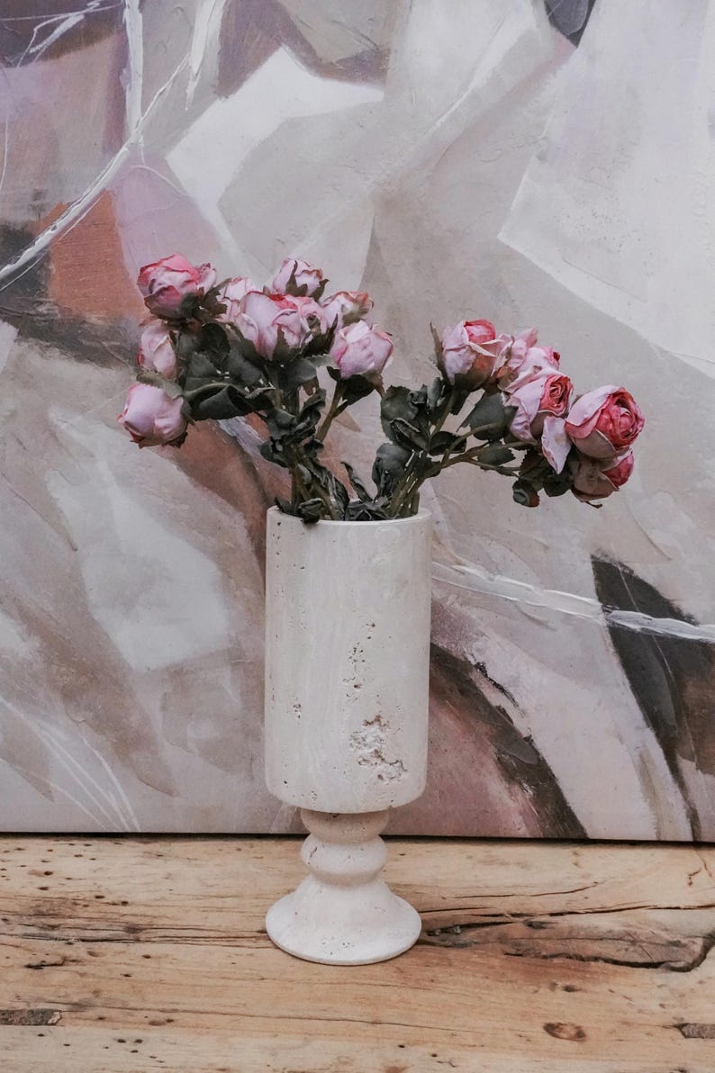 Puede incluir: Un jarr&oacute;n cil&iacute;ndrico blanco con base de pedestal, lleno de rosas rosas. El jarr&oacute;n tiene una superficie texturizada y est&aacute; sobre una superficie de madera. El fondo presenta una pintura abstracta con tonos neutros.