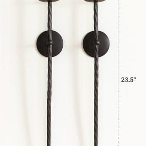 May include: Two black metal wall sconces. Each sconce has a circular base, a twisted stem, and a small, round candle holder. The sconces are 23.5 inches tall and 5 inches wide.