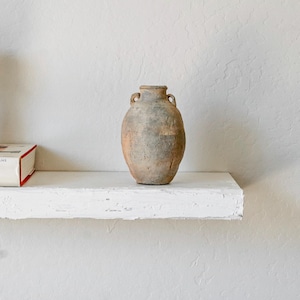 May include: A small, weathered terracotta vessel with a rounded body, a narrow neck, and two small handles on either side. The surface features a mottled, earthy texture with shades of tan, gray, and brown, showing signs of age and wear.