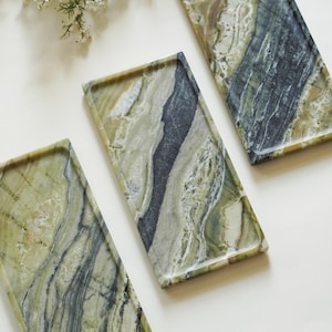 May include: Three rectangular trays crafted from polished green marble, each featuring a unique natural veining pattern with swirls of dark charcoal, sage green, and creamy white tones. The trays have slightly raised edges and a smooth finish.
