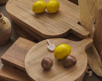 May include: Wooden cutting boards of various shapes and sizes, some with handles. Two yellow lemons rest on a rectangular board, while a round board holds a lemon, two plums, and a small white spatula. The boards are made of light brown wood.