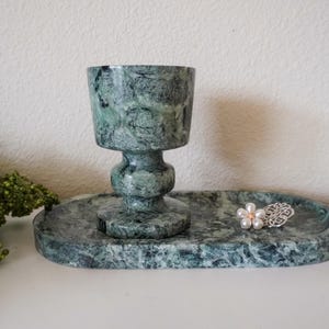 May include: A green marble chalice-shaped cup with a matching oval tray. A decorative silver and pearl flower-shaped ring sits on the tray. The set is displayed on a white surface.