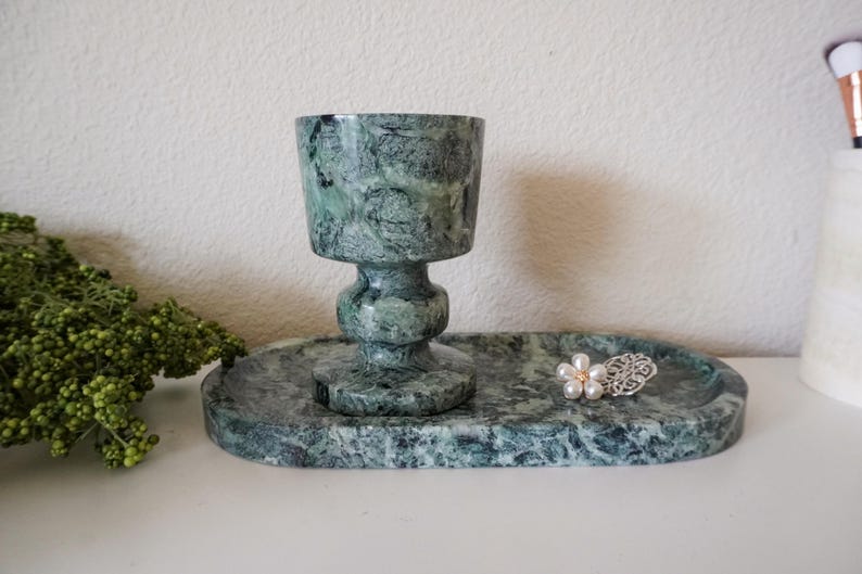 May include: A green marble chalice-shaped cup with a matching oval tray. A decorative silver and pearl flower-shaped ring sits on the tray. The set is displayed on a white surface.