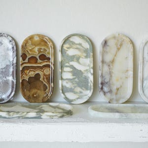May include: A collection of seven oval-shaped stone trays featuring various natural patterns and colors, including white, grey, brown, and green marble and onyx. Five trays are standing upright, while two trays are lying flat in the foreground.