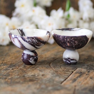 May include: Two small, bowl-shaped vessels crafted from white and dark purple marbled stone. Each bowl features a smooth, rounded interior and exterior, supported by a spherical base made of the same marbled material, creating a pedestal effect.