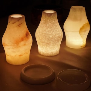 May include: Three translucent stone candle holders with internal illumination, each featuring a unique organic pattern in shades of cream, white, and amber. Two circular stone bases are positioned in front of the illuminated vessels.