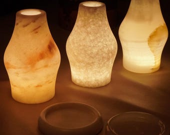 Puede incluir: Tres portavelas de piedra translúcida con iluminación interna, cada uno con un patrón orgánico único en tonos crema, blanco y ámbar. Dos bases circulares de piedra están colocadas frente a los recipientes iluminados.