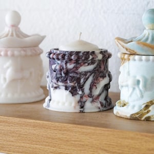May include: Three cylindrical candle holders with carousel-inspired relief designs. The center holder features a white and dark purple marbled pattern with a white wax candle insert. The flanking holders are white and pastel-toned with lids.