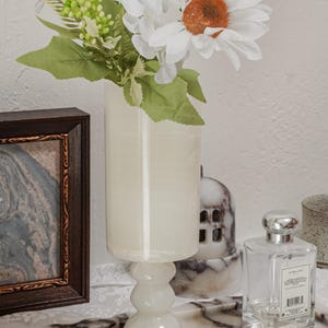 Puede incluir: Un jarr&oacute;n blanco con base de pedestal contiene flores artificiales blancas y verdes. Una foto enmarcada, un frasco de perfume y una vela se encuentran en una bandeja con estampado de m&aacute;rmol. Tambi&eacute;n hay anillos de oro en la bandeja.