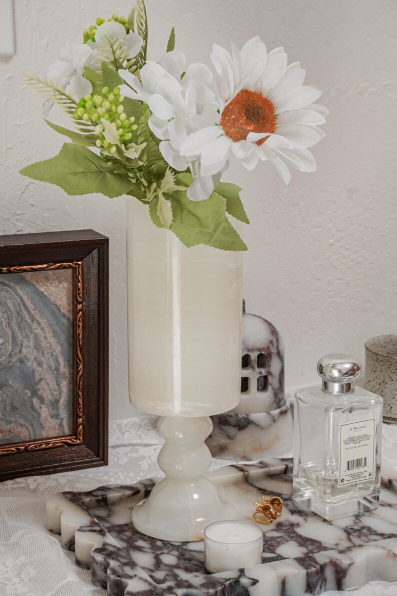 Puede incluir: Un jarr&oacute;n blanco con base de pedestal contiene flores artificiales blancas y verdes. Una foto enmarcada, un frasco de perfume y una vela se encuentran en una bandeja con estampado de m&aacute;rmol. Tambi&eacute;n hay anillos de oro en la bandeja.