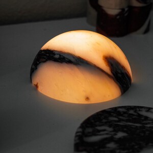 Puede incluir: Una l&aacute;mpara en forma de c&uacute;pula hecha de piedra transl&uacute;cida con una luz c&aacute;lida y brillante. La piedra presenta vetas naturales en tonos marr&oacute;n oscuro y negro sobre una base de color blanco crema. La luz emana de la superficie curva.