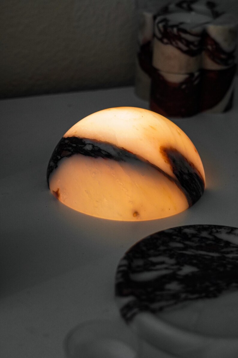 Puede incluir: Una l&aacute;mpara en forma de c&uacute;pula hecha de piedra transl&uacute;cida con una luz c&aacute;lida y brillante. La piedra presenta vetas naturales en tonos marr&oacute;n oscuro y negro sobre una base de color blanco crema. La luz emana de la superficie curva.